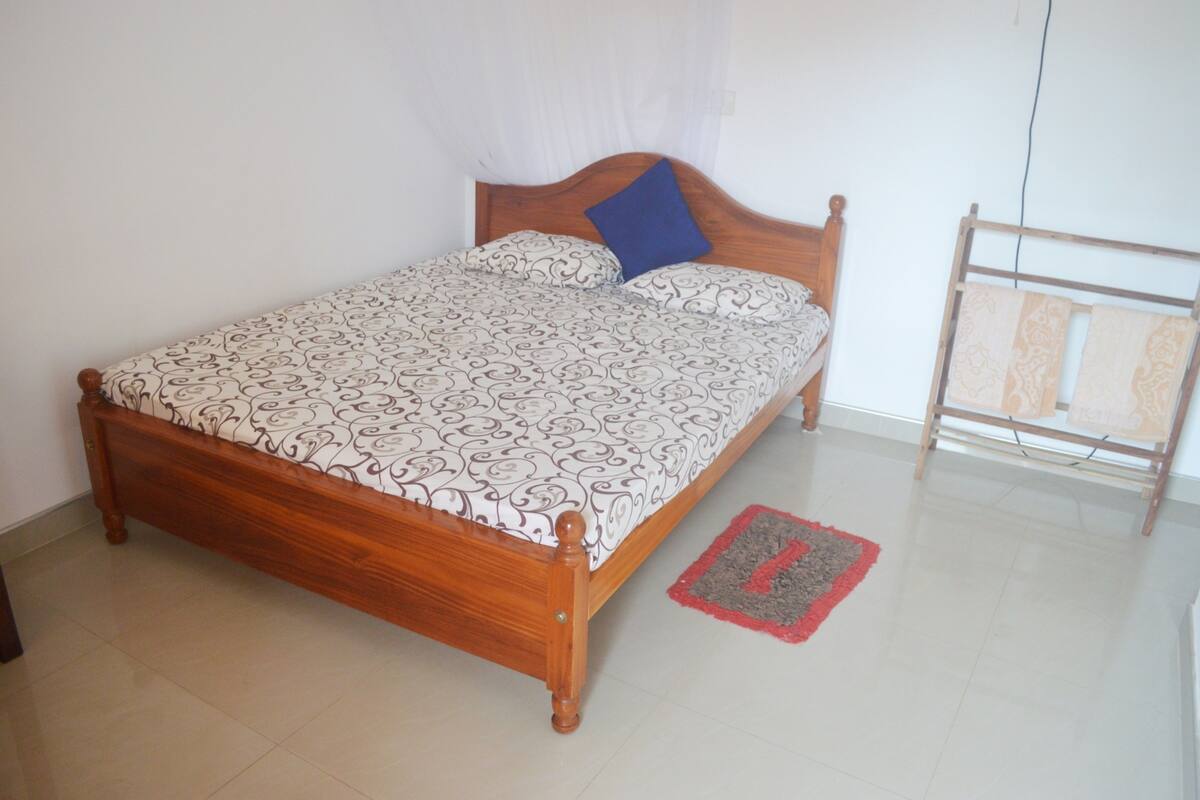 Un lit simple avec une couvre-lit à motifs et un oreiller bleu, une tête de lit en bois et un tapis rouge et gris sur un sol carrelé.