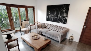Sala de estar com sofá bege, mesa de centro de madeira, uma cadeira e uma janela grande com vista para área verde.