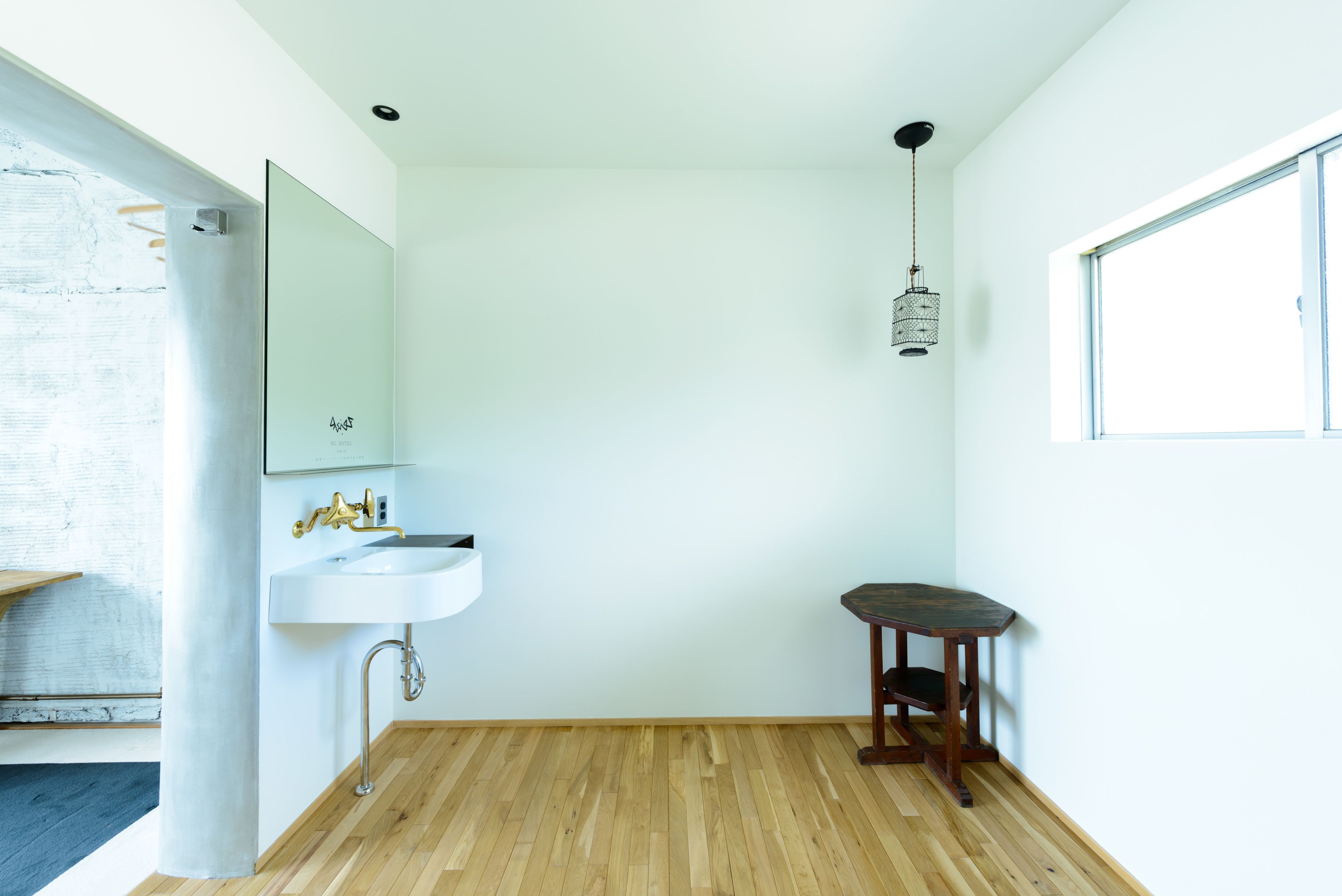 Un baño minimalista con un lavamanos blanco, suelo de madera, una mesita pequeña y una lámpara colgante.