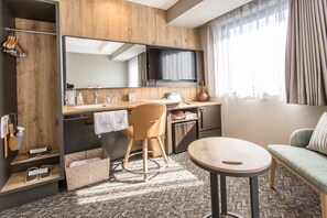 A modern hotel room with a wooden accent wall, a large mirror, a flat-screen TV, a desk with a chair, a small round table, a sofa, and a window with curtains.