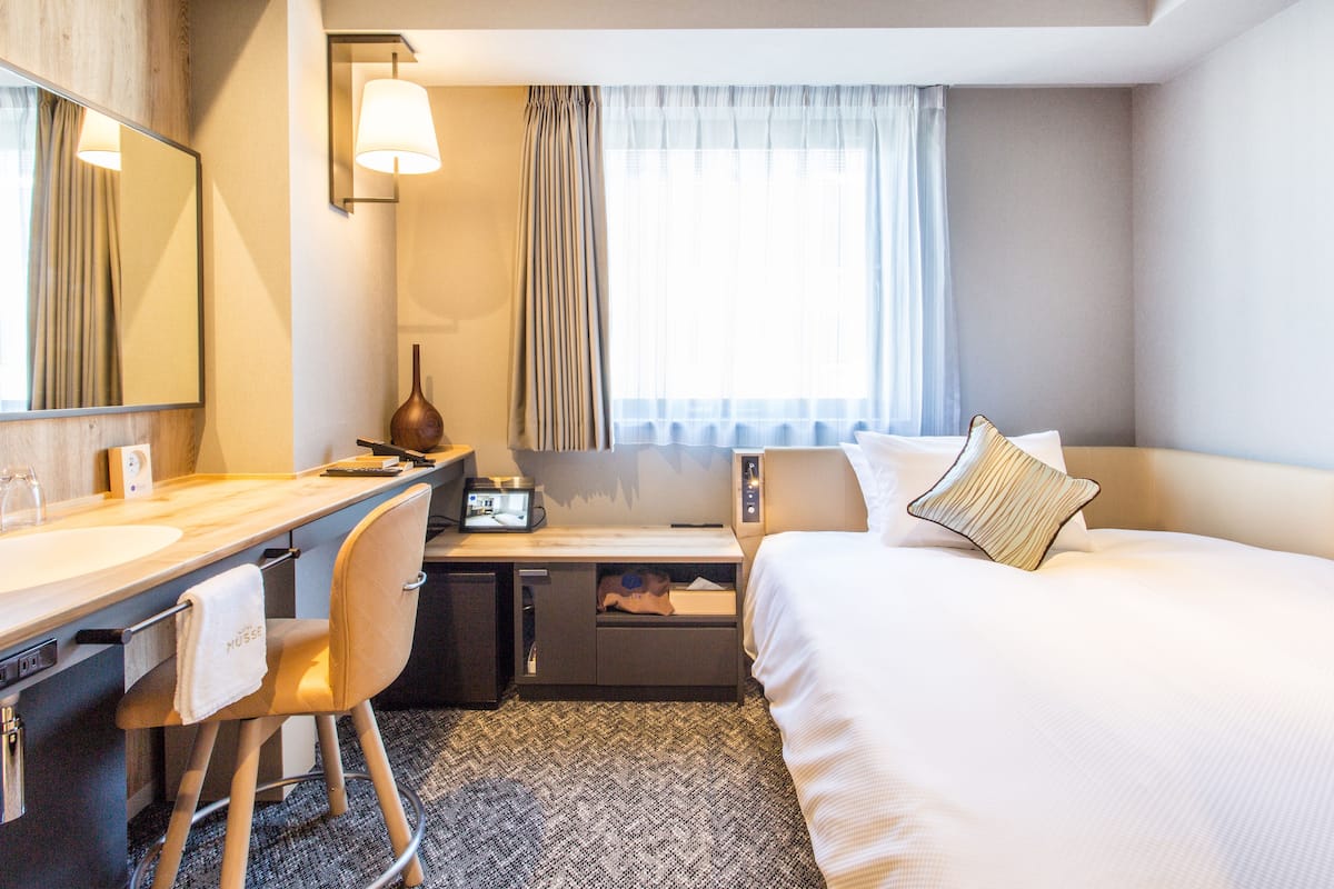 Une chambre d’hôtel comprenant un lit, un bureau avec une chaise, un ordinateur, une lampe et une fenêtre avec des rideaux.