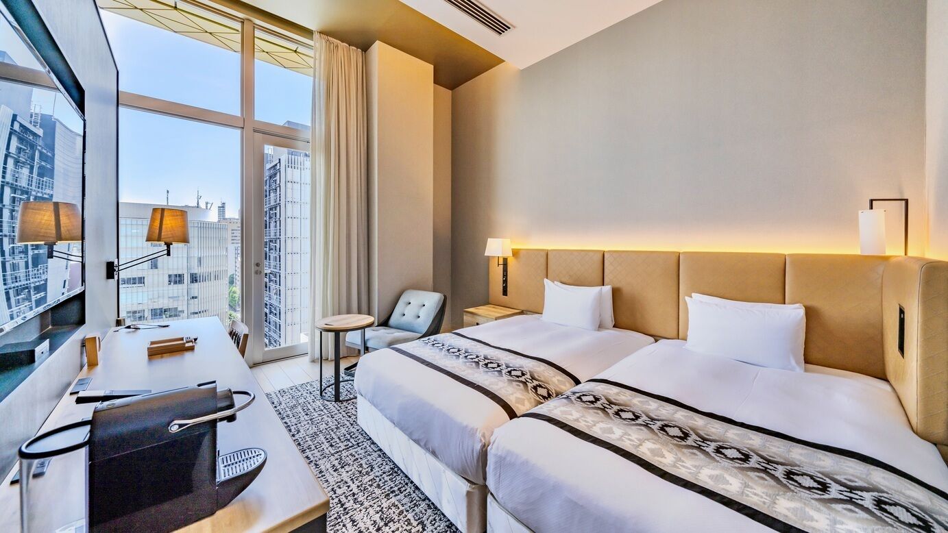 A hotel room with two beds, a desk, a chair, and a large window offering a city view.