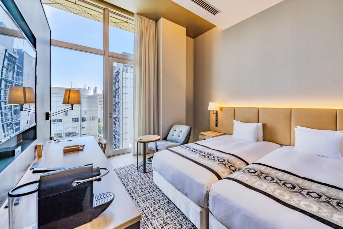 Ein Hotelzimmer mit zwei Betten, einem Schreibtisch, einem Sessel und einem großen Fenster mit Blick auf die Stadt.