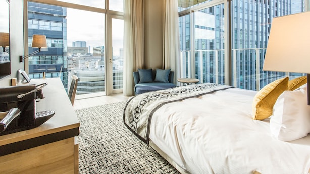 Ein Hotelzimmer mit einem großen Bett, einem Schreibtisch mit Telefon, einem kleinen Tisch und Blick auf die Stadt durch große Fenster.