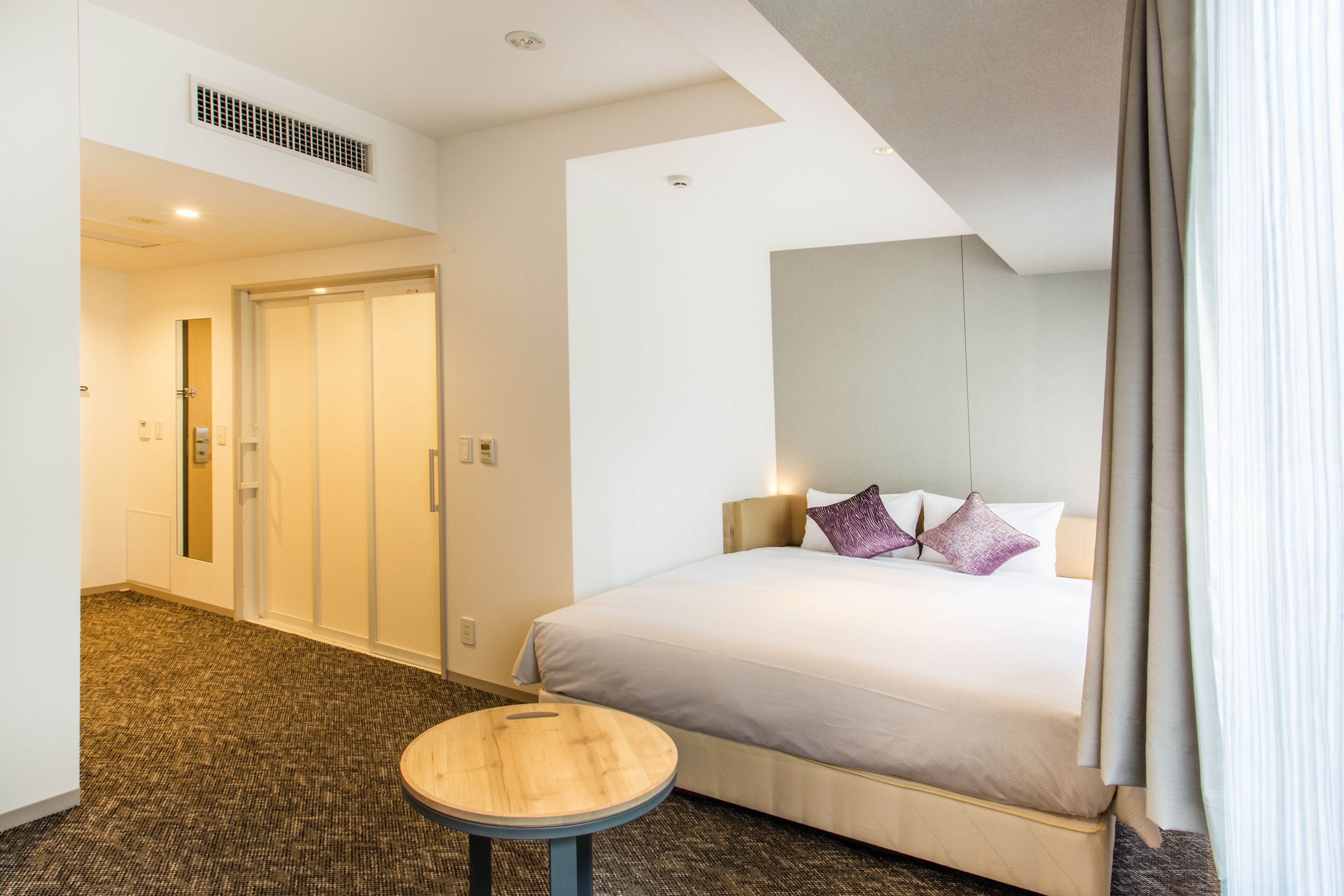 A hotel room with a bed, a round wooden table, a mirror, and a door leading to another room.