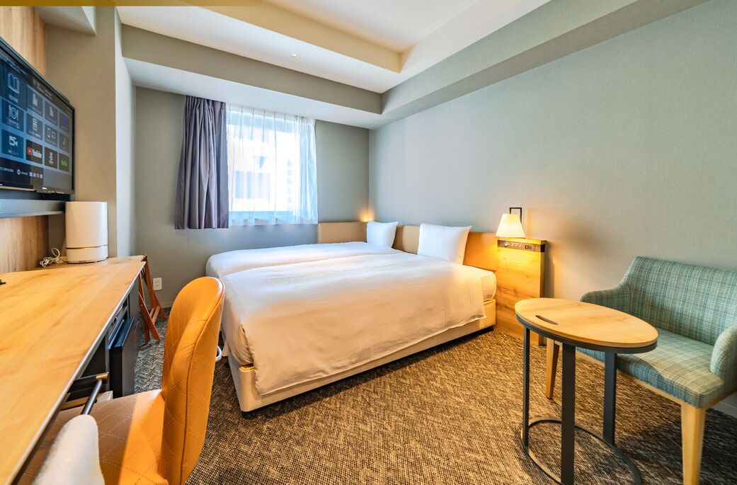 A hotel room with a bed, a desk, a chair, a small table, and a TV.