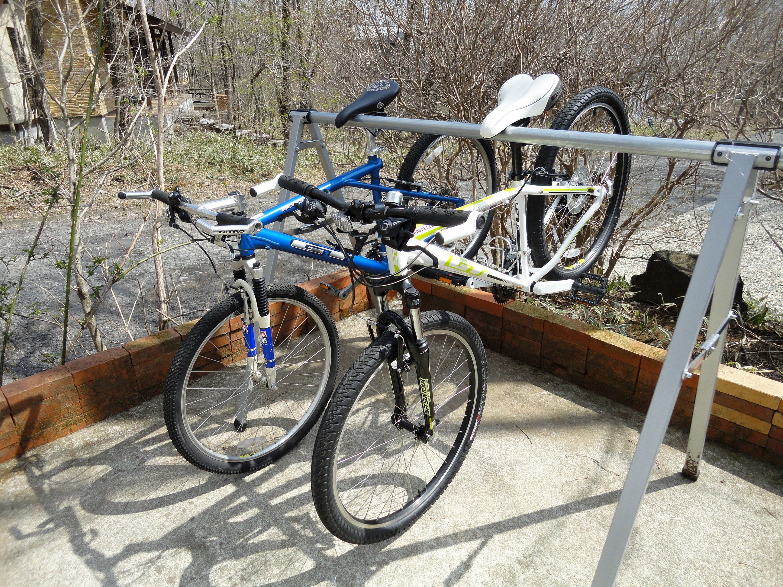 A bicycle rack holding two bicycles, one blue and one white, with a brick wall and trees in the background.