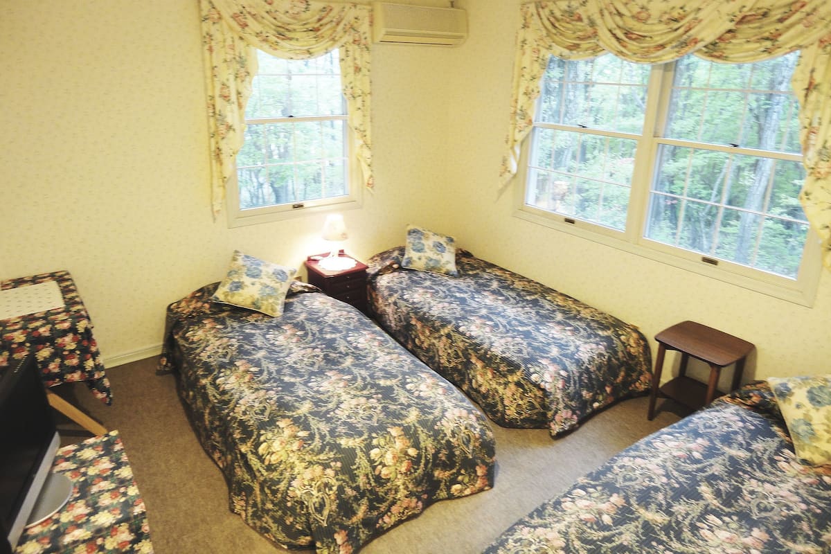 A room with two beds, a desk, and a chair. There are two windows with curtains.