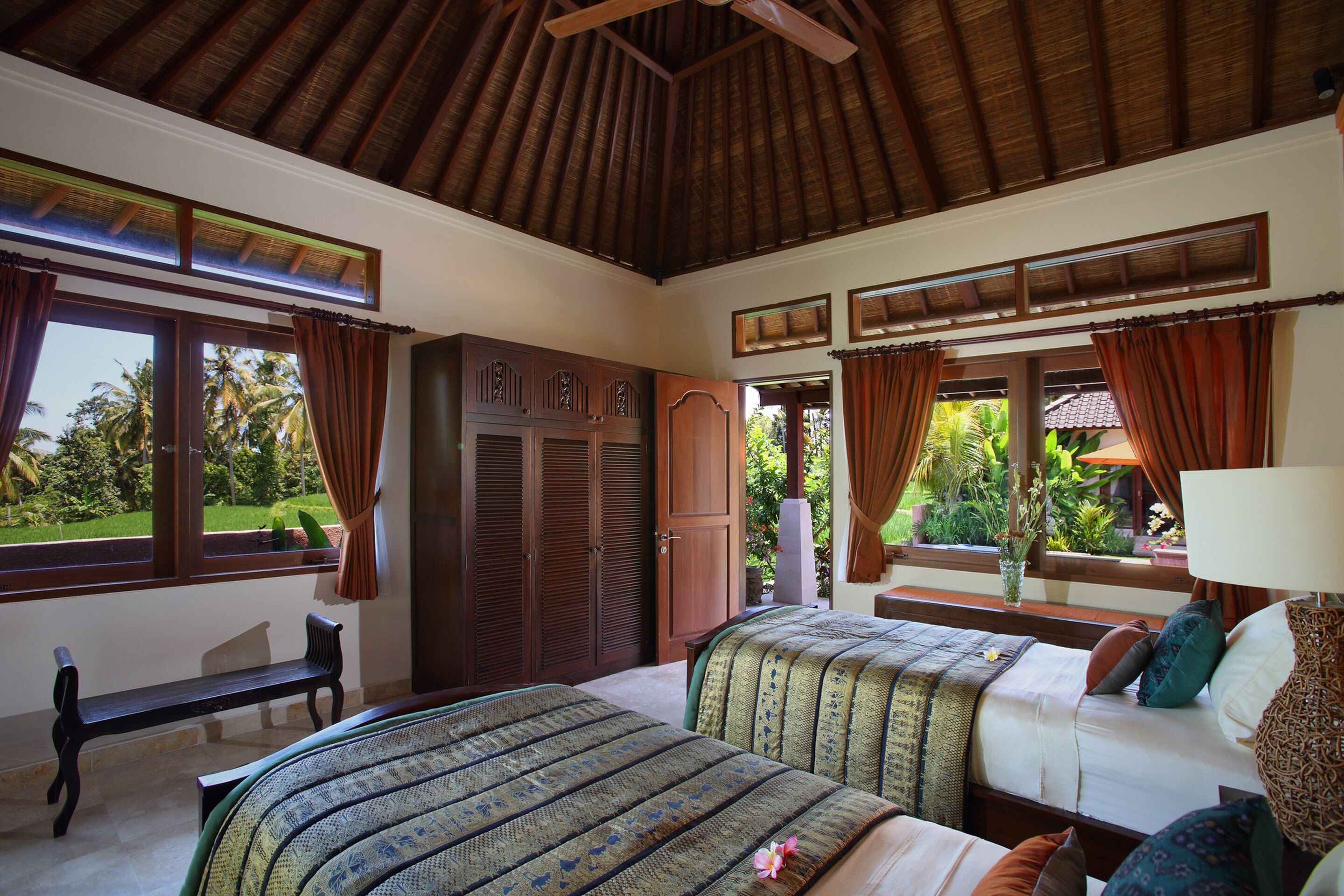 A bedroom with two beds, a bench, and a view of the garden.