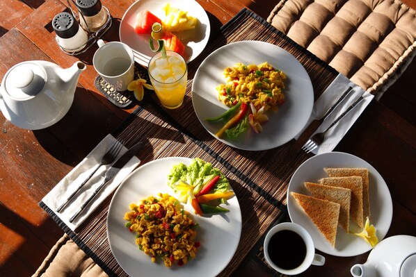 A breakfast setting with plates of scrambled eggs, toast, and fruit, along with a teapot, cups, and cutlery on a wooden table. - Satori Villas Bali (Ubud)