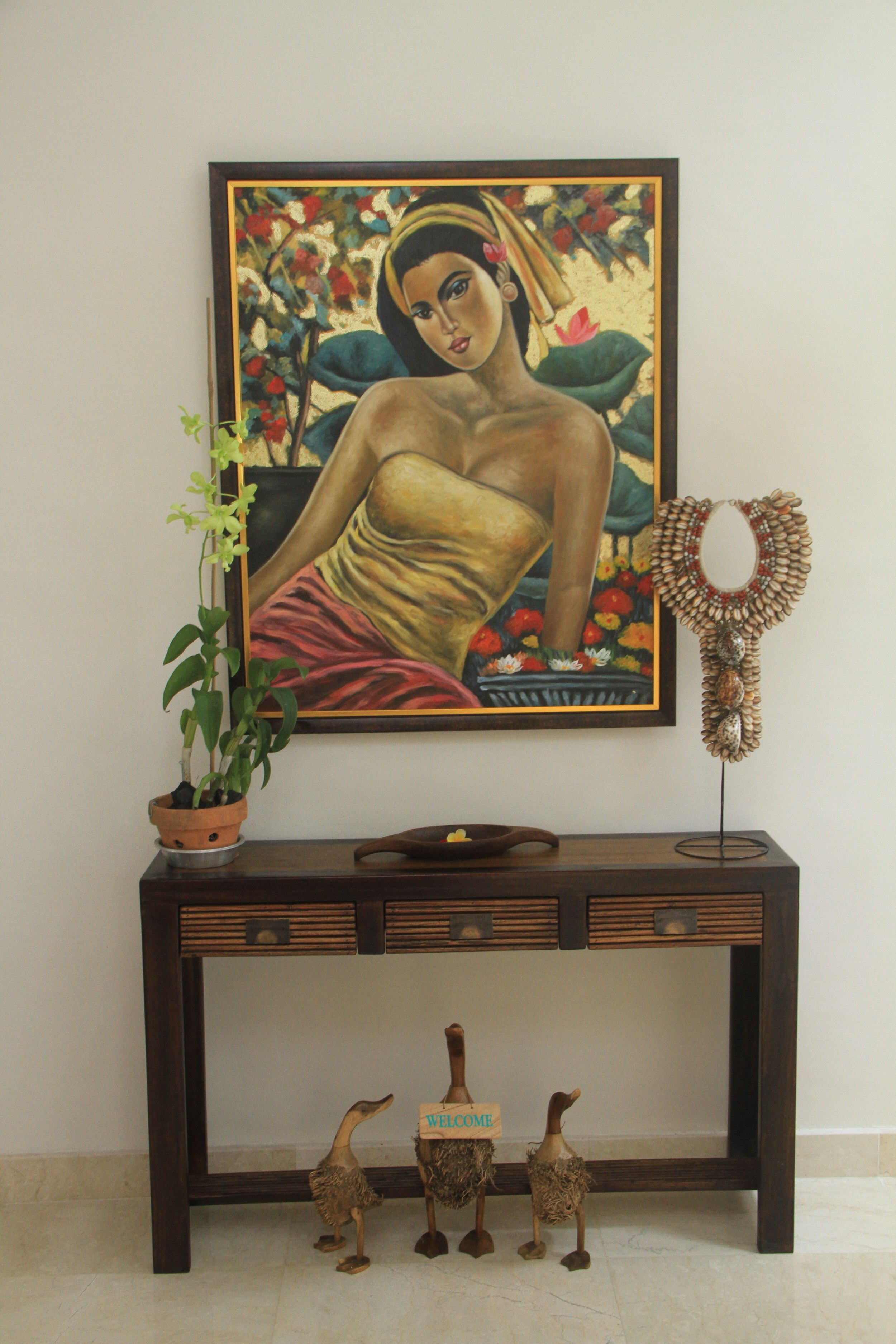 A painting of a woman on a wall, a potted plant, a decorative mirror, and a console table with figurines.