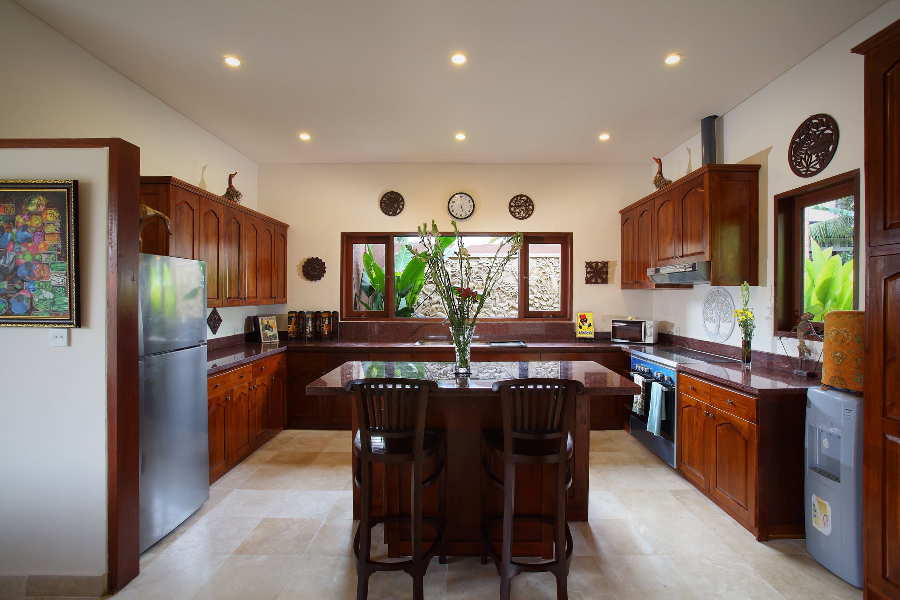A modern kitchen with wooden cabinets, a stainless steel refrigerator, a dining area with two chairs, and a large window with a view of greenery.