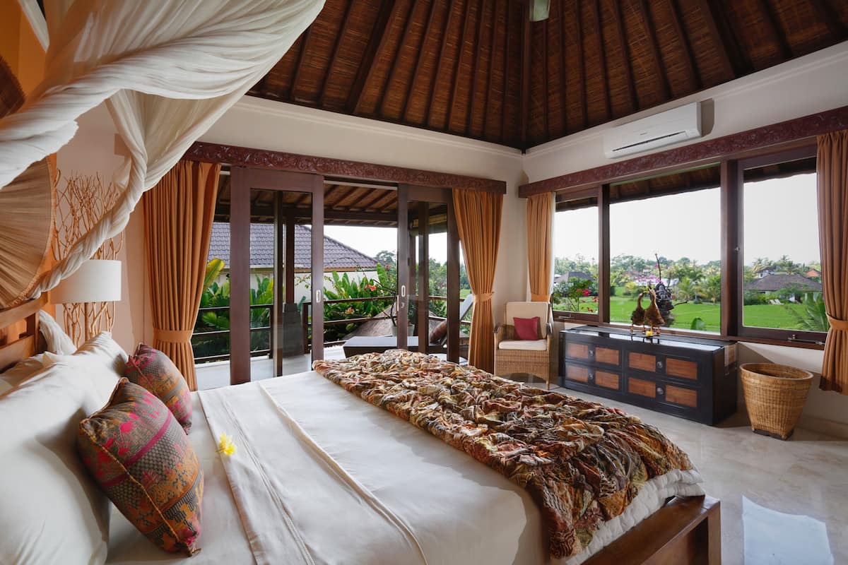 A spacious bedroom with a large bed, a nightstand, a wicker chair, and a view of the garden through large windows.