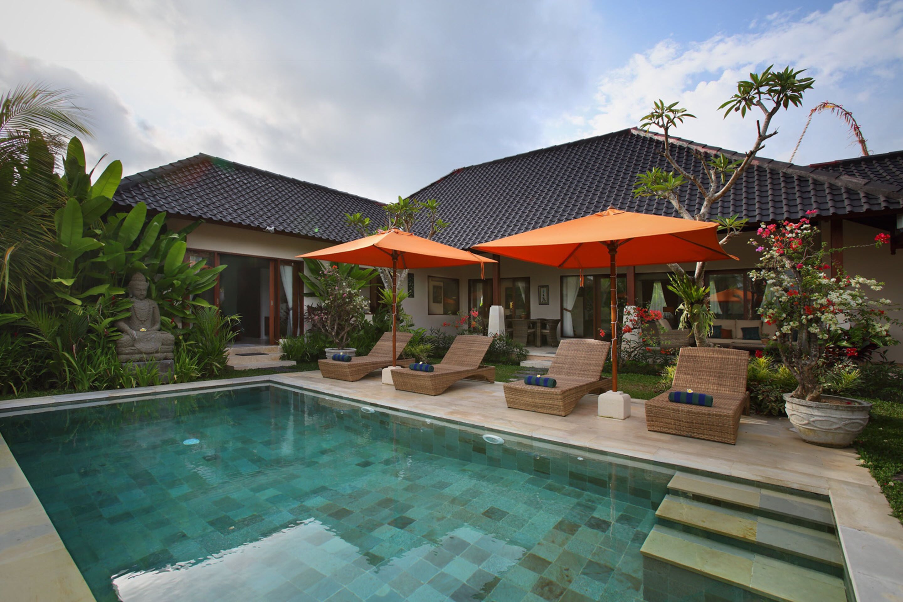 A poolside area with lounge chairs and umbrellas, surrounded by tropical plants and a tiled roof.