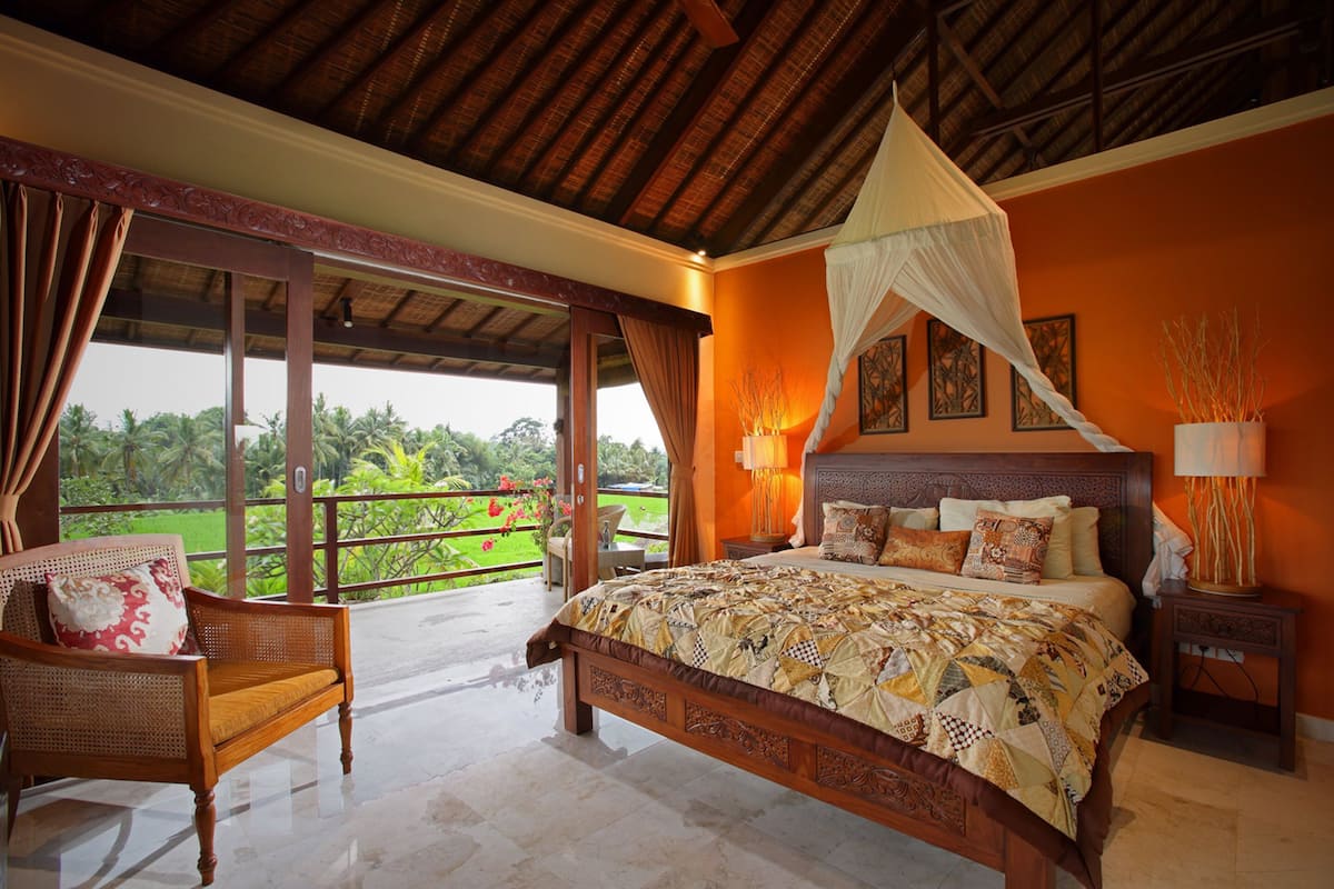 A bedroom with a large bed, a nightstand, a chair, and a view of a lush garden.