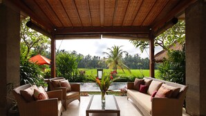 Un espacio al aire libre techado con mobiliario de mimbre y una mesa con superficie de vidrio, con vistas a un jardín exuberante y una fuente de agua.