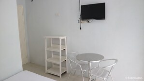 A small room with a white wall, a white shelf unit, a round table with two chairs, and a mounted TV. - Oceana Lodge (Baler)
