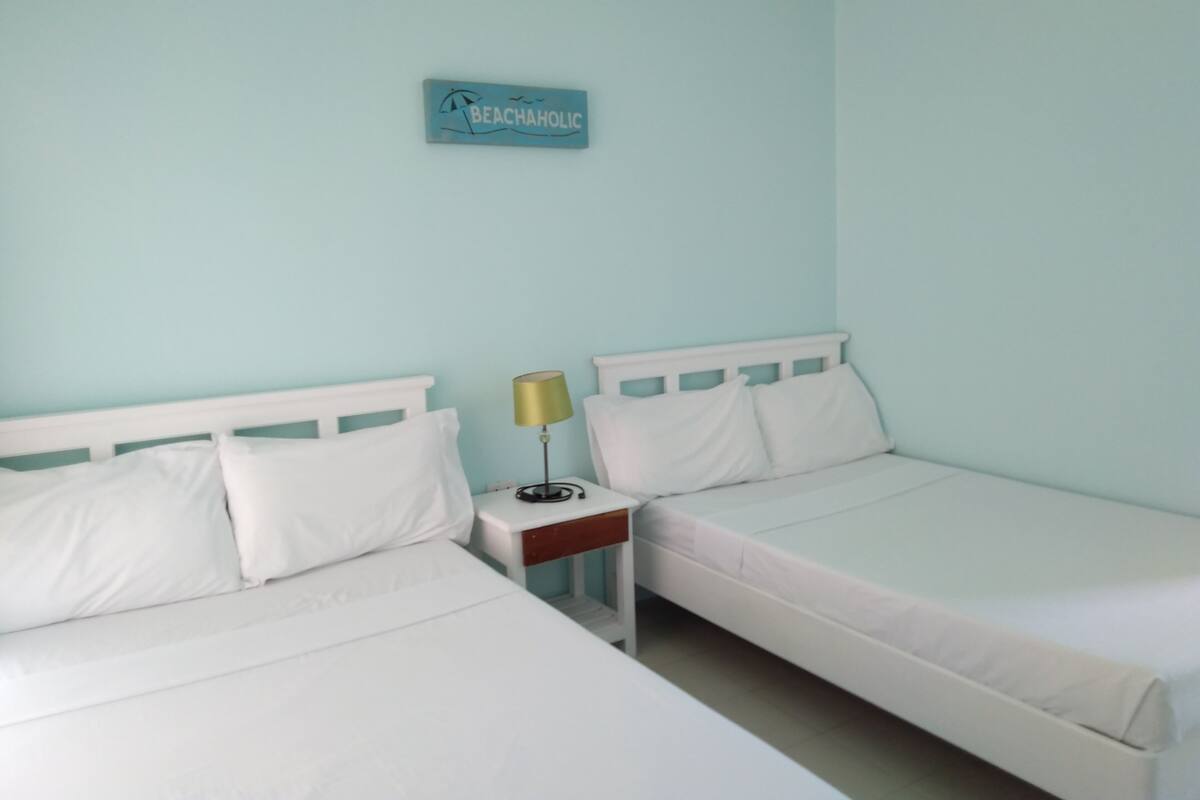 A room with two single beds, a nightstand with a lamp, and a wall sign that reads "BEACHAHOLIC".