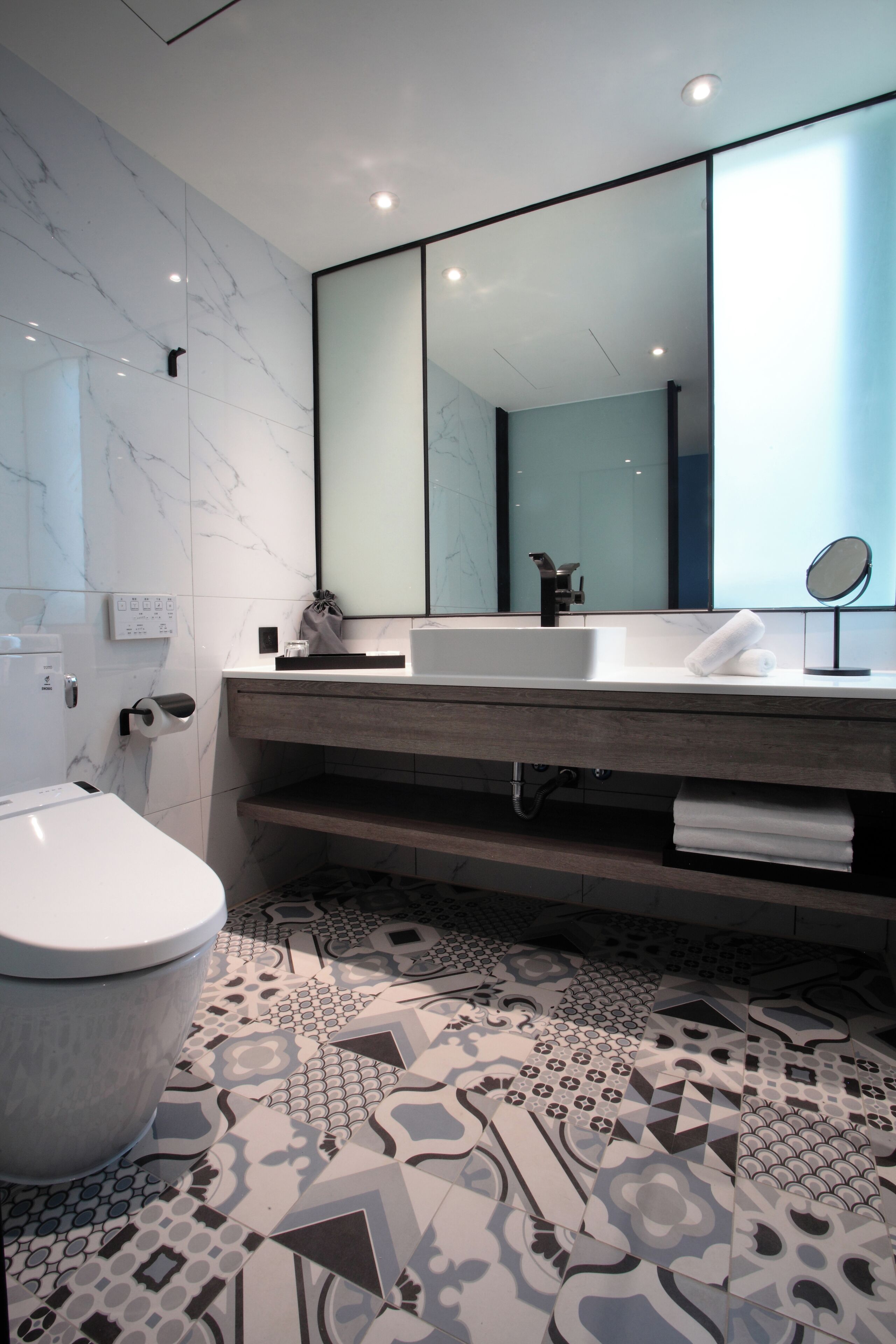 A modern bathroom with a marble wall, a large mirror, a white sink, and a patterned floor.