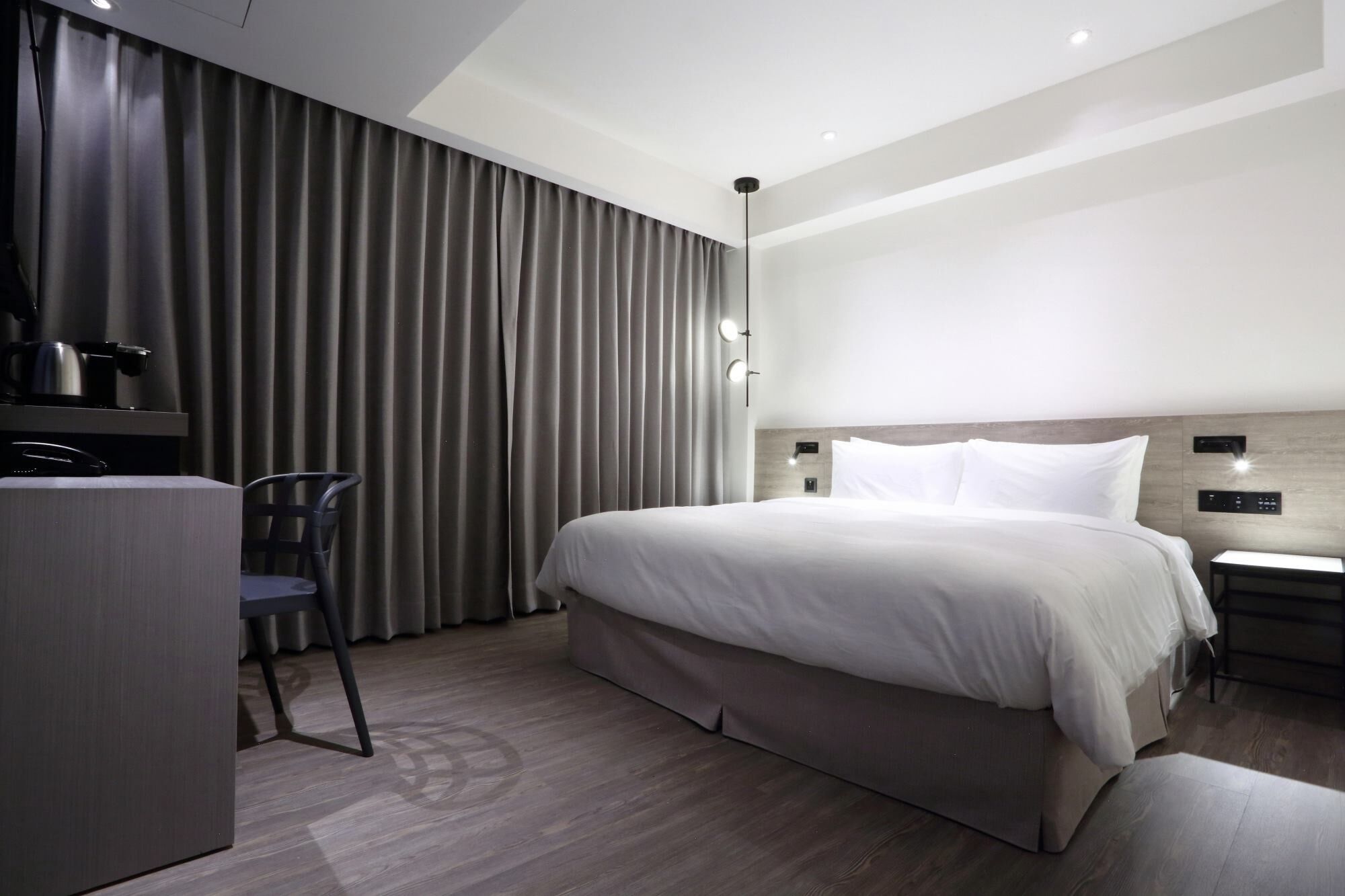 A modern hotel room with a large bed, a desk with a chair, a nightstand, and a wall-mounted light fixture.