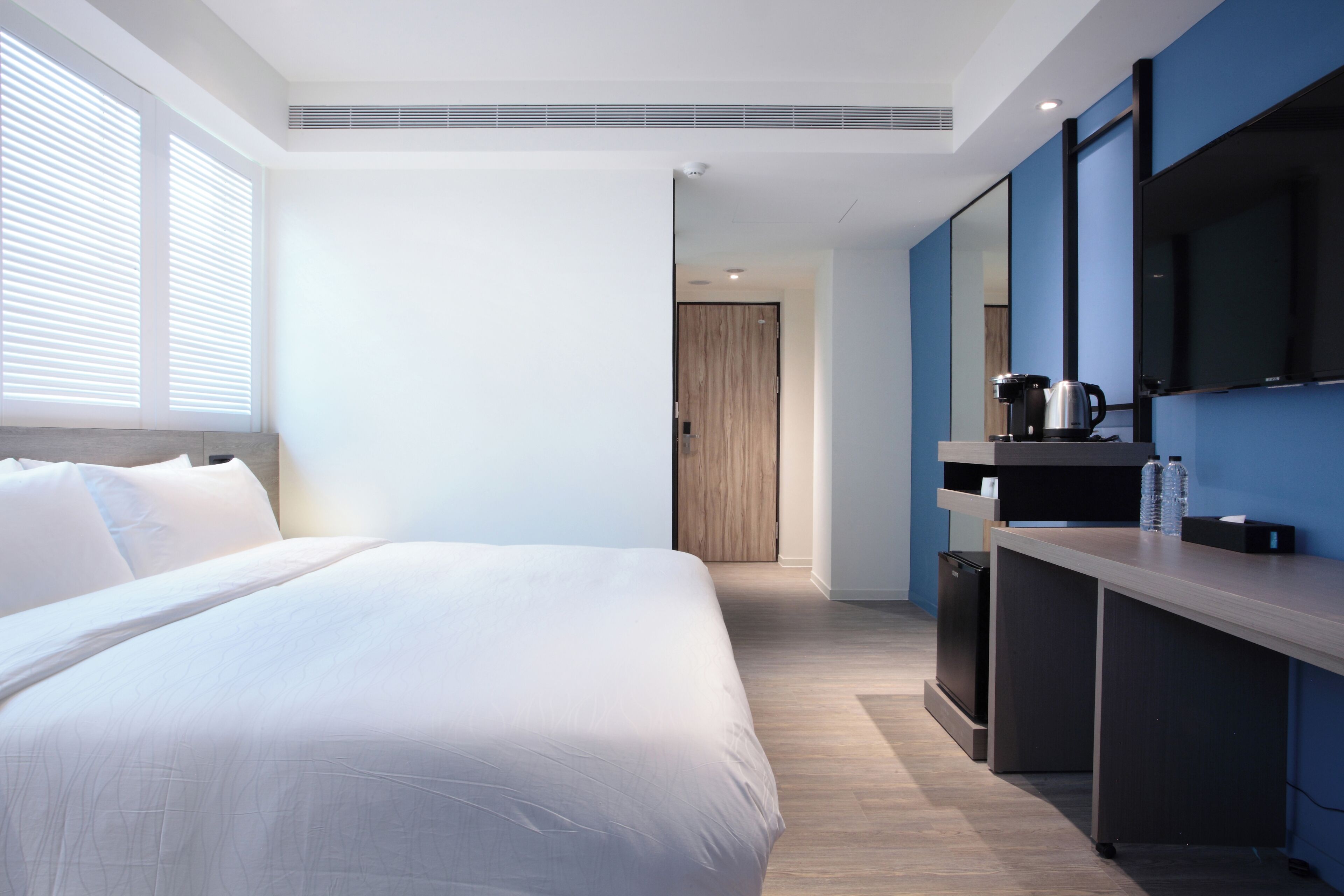 A modern hotel room with a large bed, a desk with a kettle, and a television.