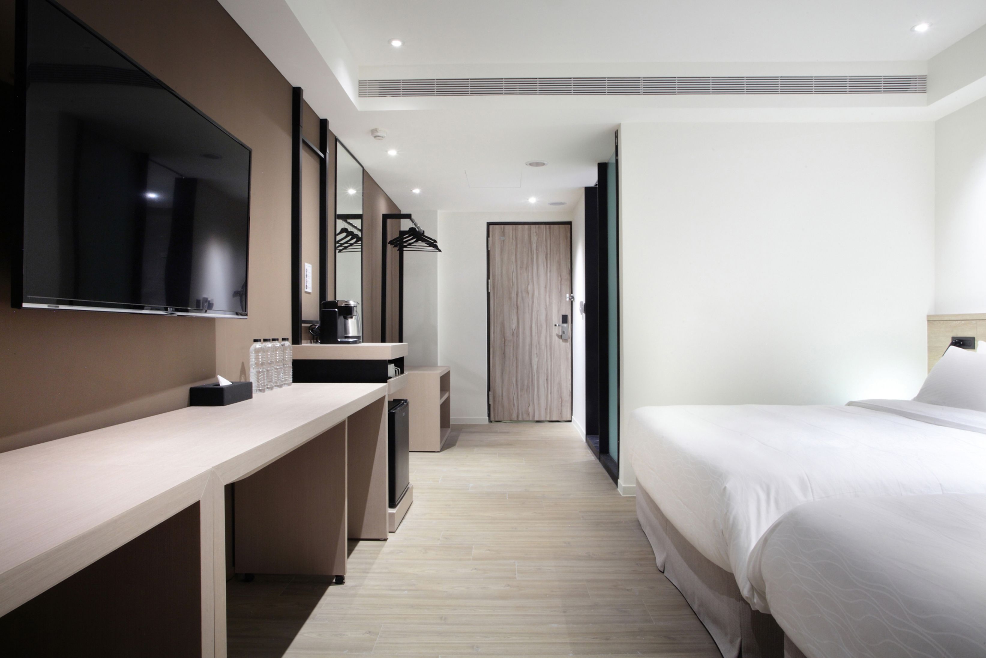 A modern hotel room with a large flat-screen TV, a desk with a coffee machine, a wardrobe, and a bed with white linens.