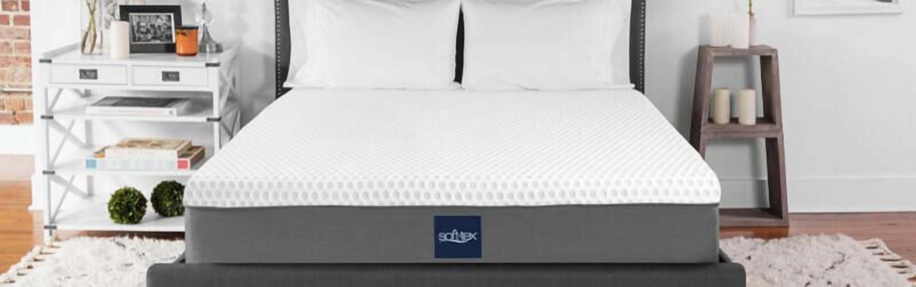 Un lit avec un matelas blanc et une base grise, une table de chevet blanche avec une lampe, et une petite étagère avec des livres et des plantes.