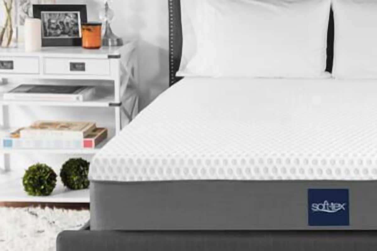 Una cama con colchón blanco y base gris, una mesita de noche blanca con una lámpara, y una pequeña estantería con libros y plantas.