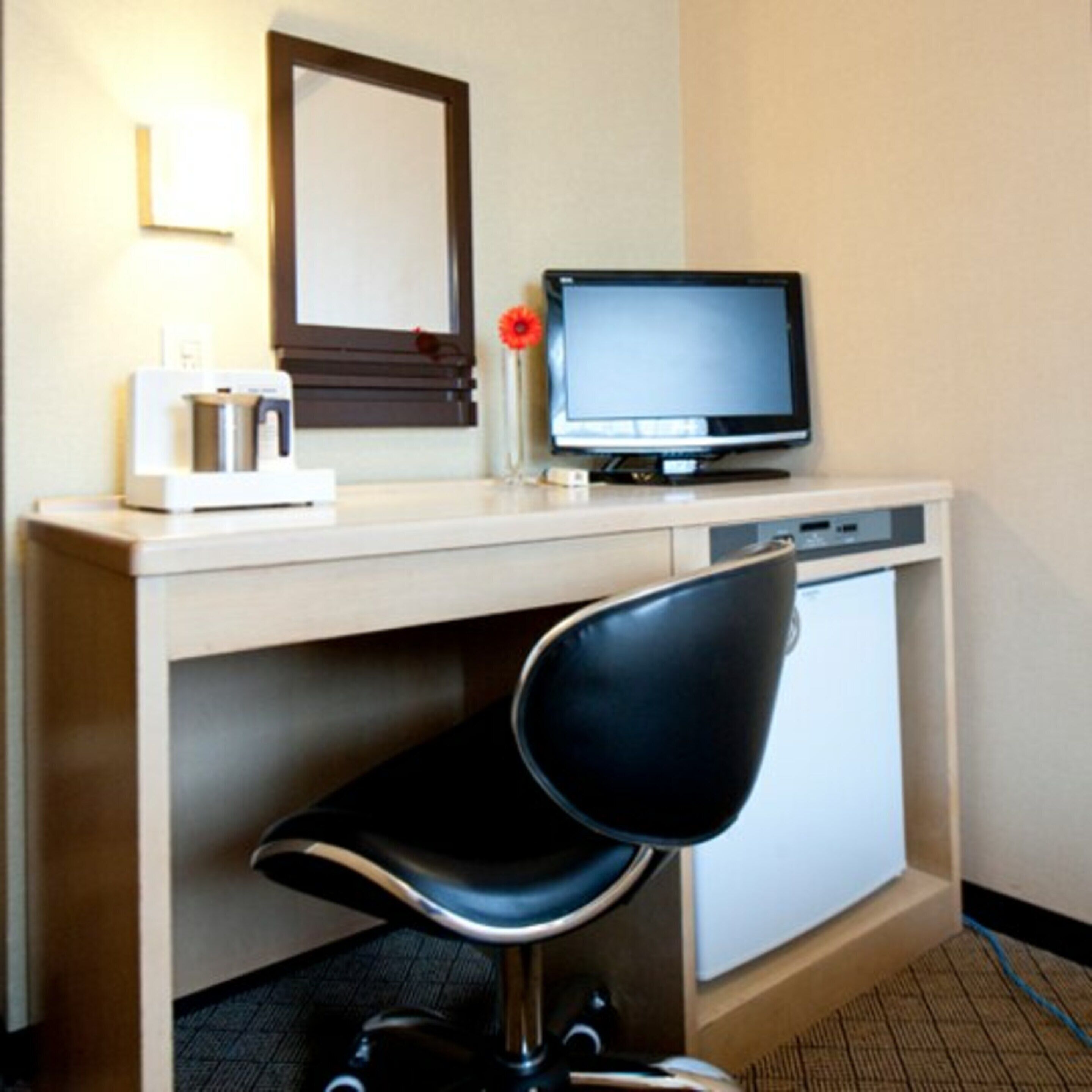 A hotel room with a desk, a computer, a chair, and a small refrigerator.