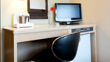 A hotel room with a desk, a computer, a chair, and a small refrigerator.