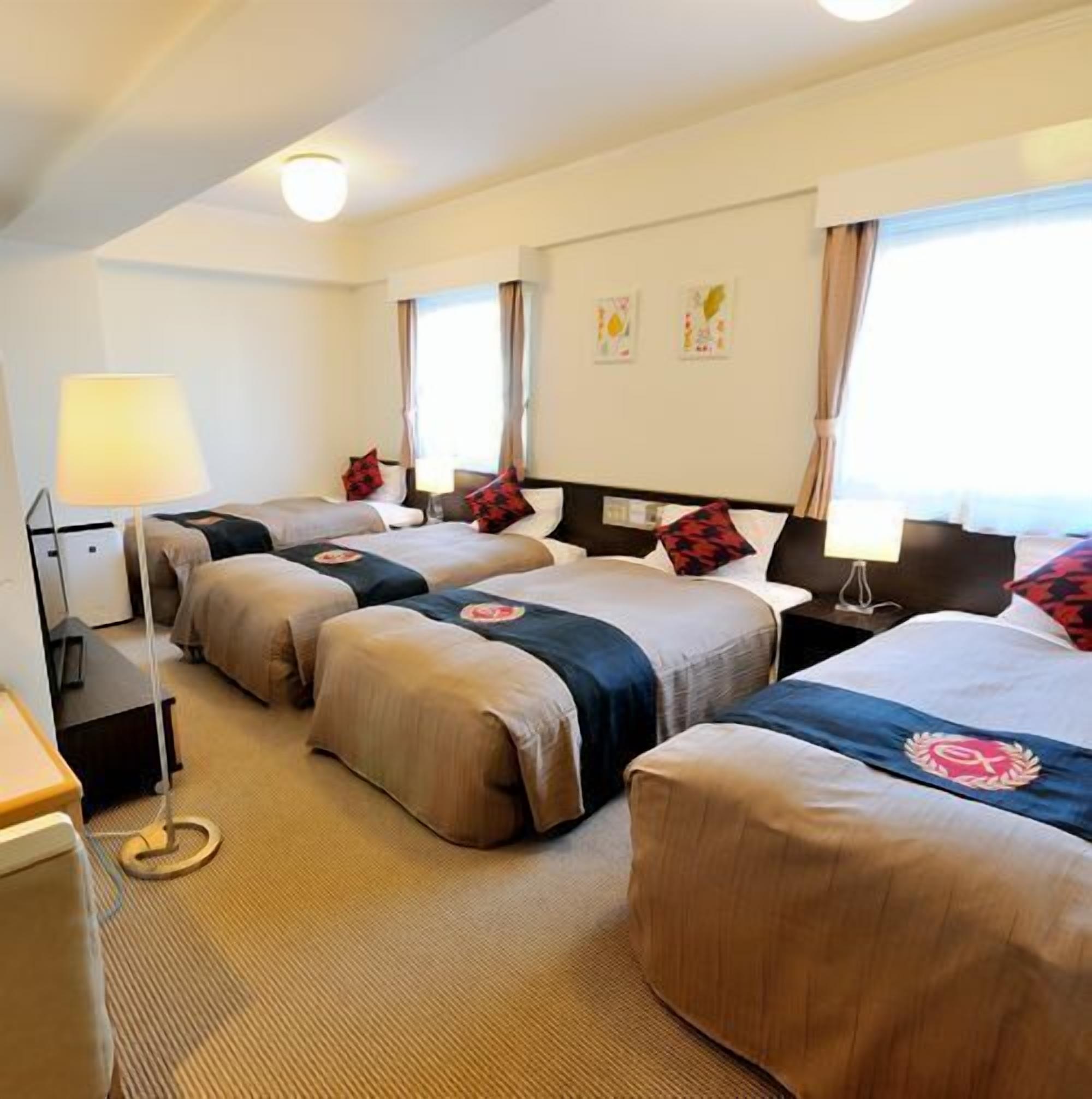 Une chambre d’hôtel avec quatre lits disposés en rangée, un bureau avec une lampe et une fenêtre avec des rideaux.