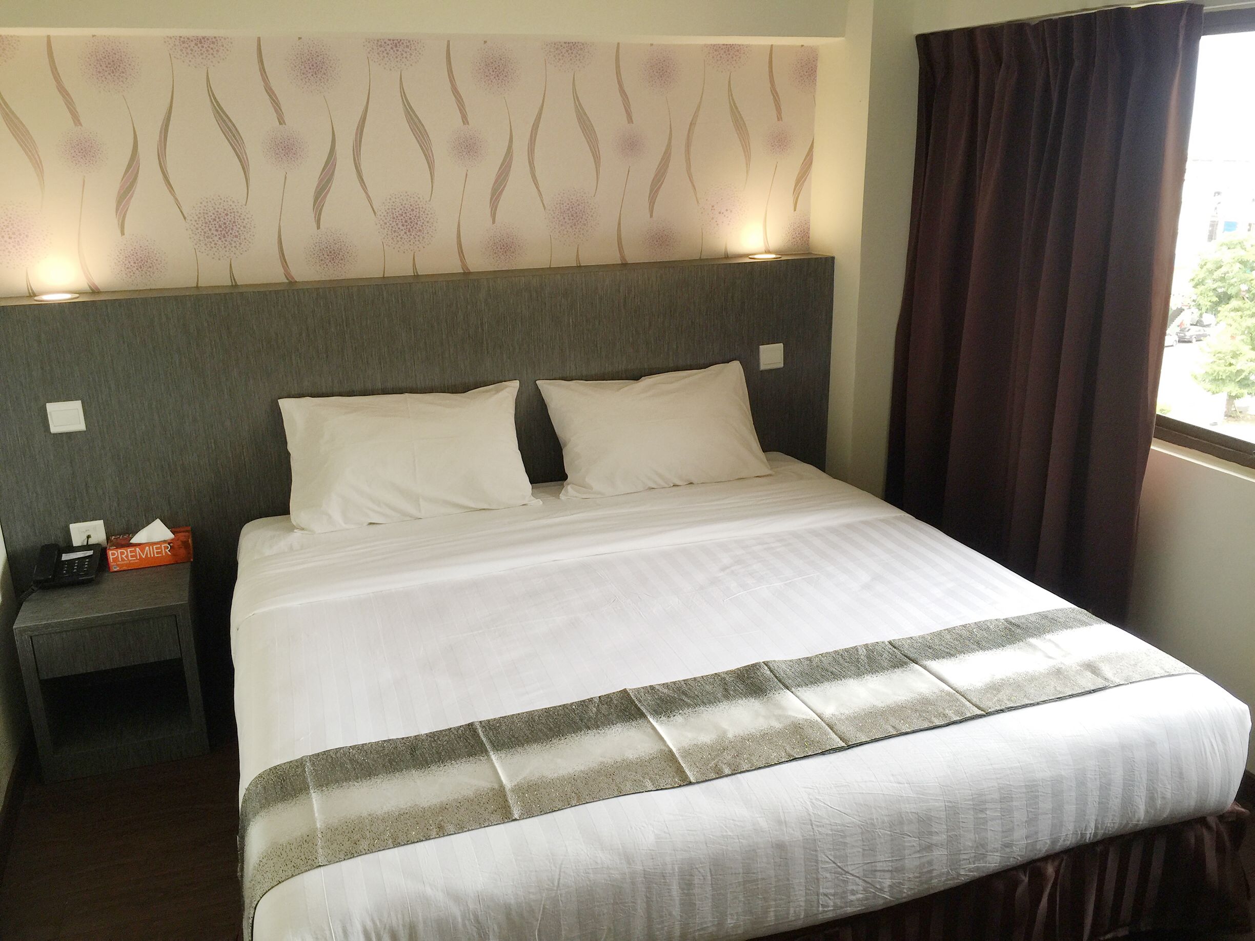 Una cama bien hecha con ropa blanca y una franja gris decorativa, una mesita de noche con un teléfono y una caja de pañuelos, y una pared con estampado floral.