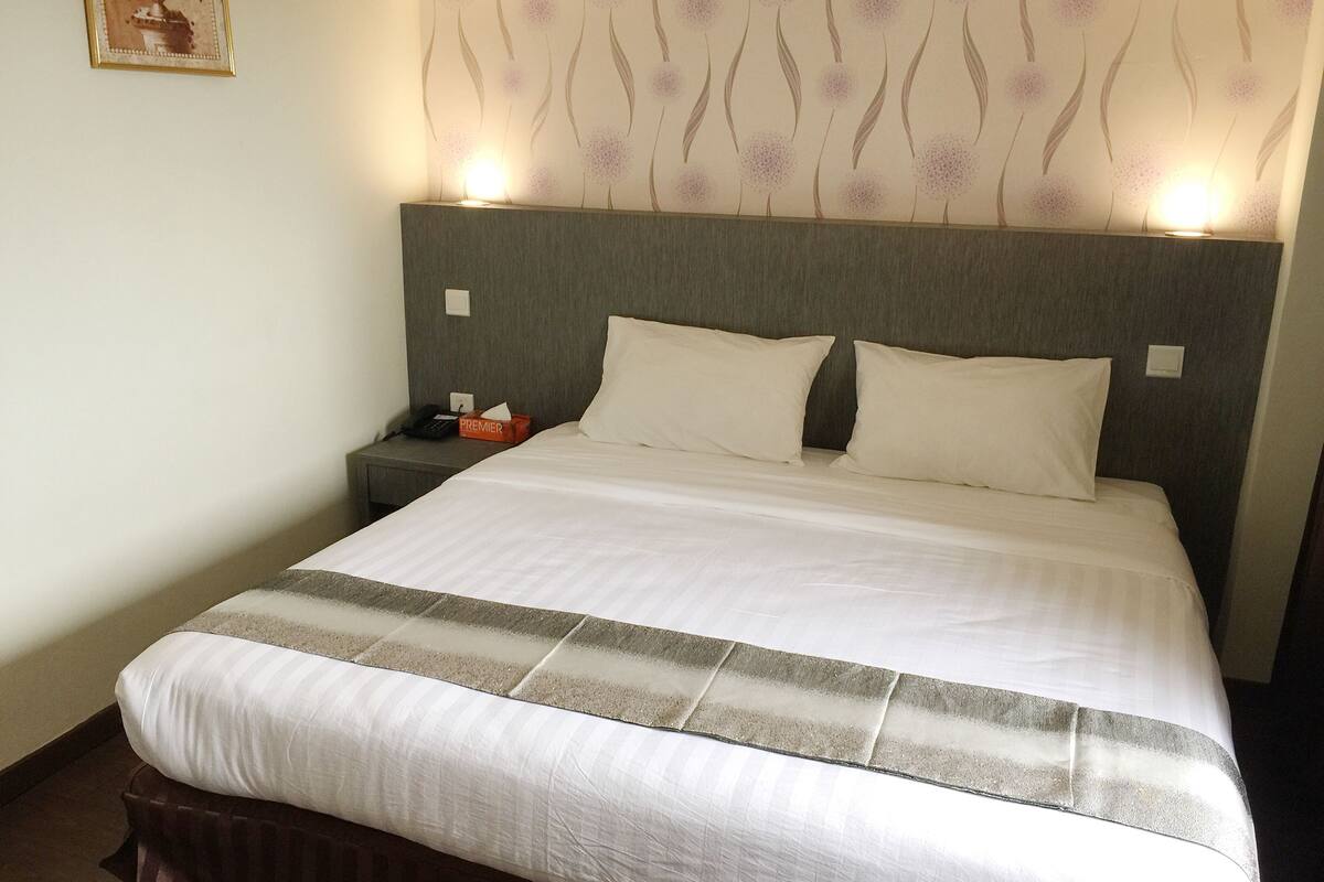 A neatly made bed with white linens and a grey headboard, a bedside table with a lamp, and a wall with a floral wallpaper pattern.