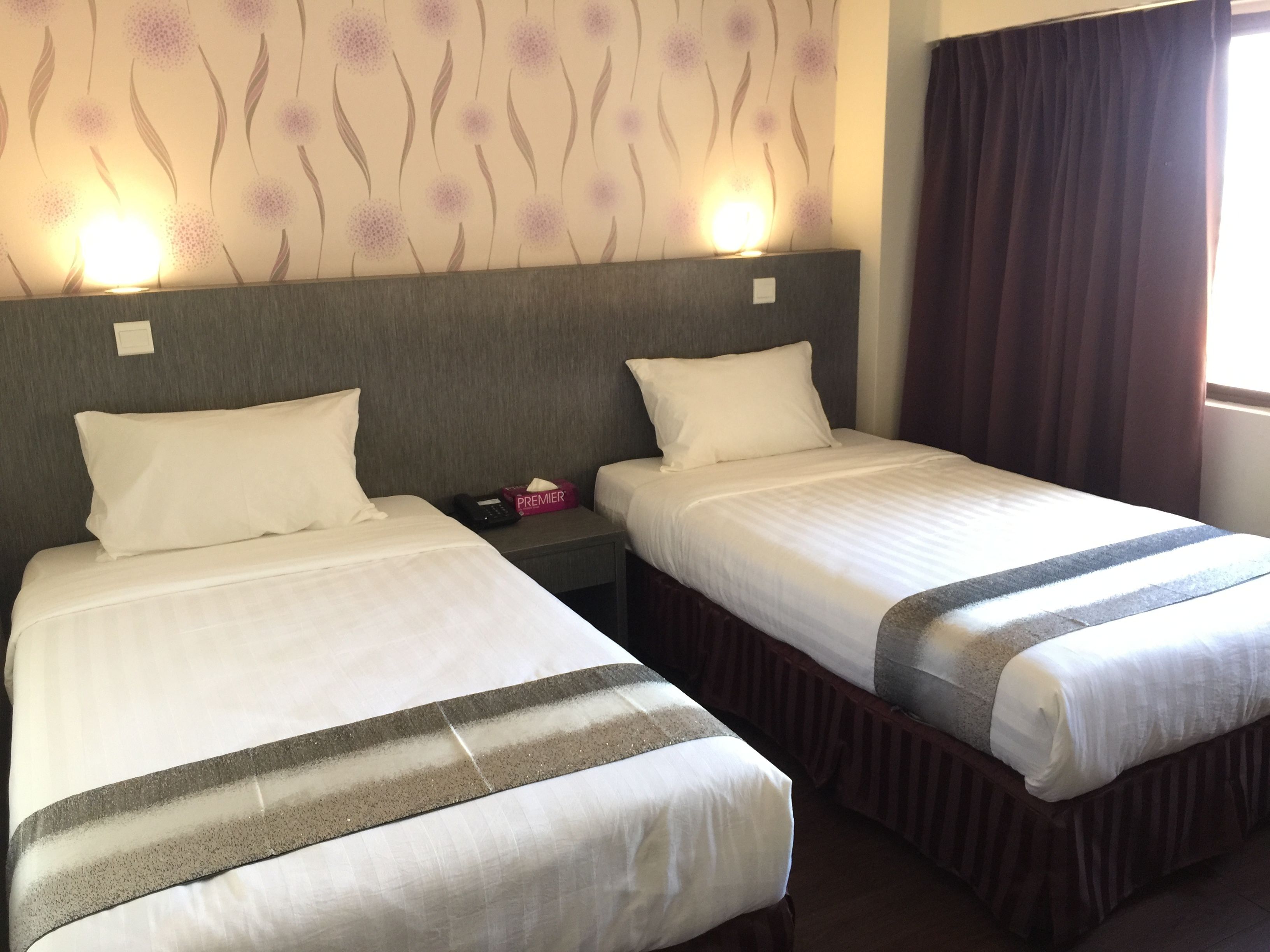 Two single beds with white linens and a grey headboard, a bedside table with a phone and a tissue box, and a wall with a floral pattern.