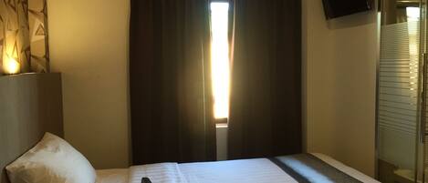 A hotel room with a bed, a bedside table with a phone and remote, a TV on the wall, and a window with curtains.