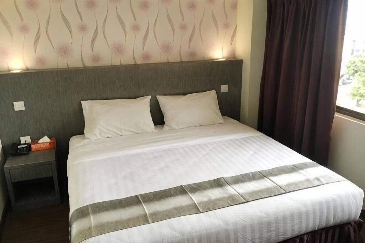 A hotel room with a bed, a nightstand, a window with curtains, and a wall with a floral pattern.
