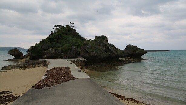 Ein Küstenweg, der zu einer felsigen Insel mit Sandstrand und ruhiger See führt.