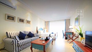 Quarto de hotel com sofá listrado, mesa de centro de madeira, TV em suporte de madeira e cama com cabeceira azul.