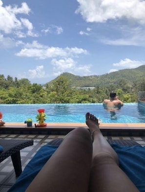 A poolside view with a person relaxing on a lounge chair, another person swimming in the pool, and a view of a lush green landscape in the background.