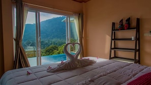 A hotel room with a bed, a large window with curtains, a view of a mountain and a pool, and a wooden shelf with decorative items.