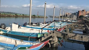 Una marina con varias embarcaciones amarradas, un muelle con barandilla de metal y un paisaje urbano al fondo.