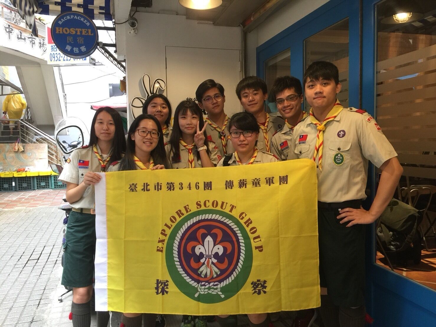 A group of individuals in scout uniforms holding a yellow banner with a logo and text, standing in an indoor setting with a blue wall and a window.