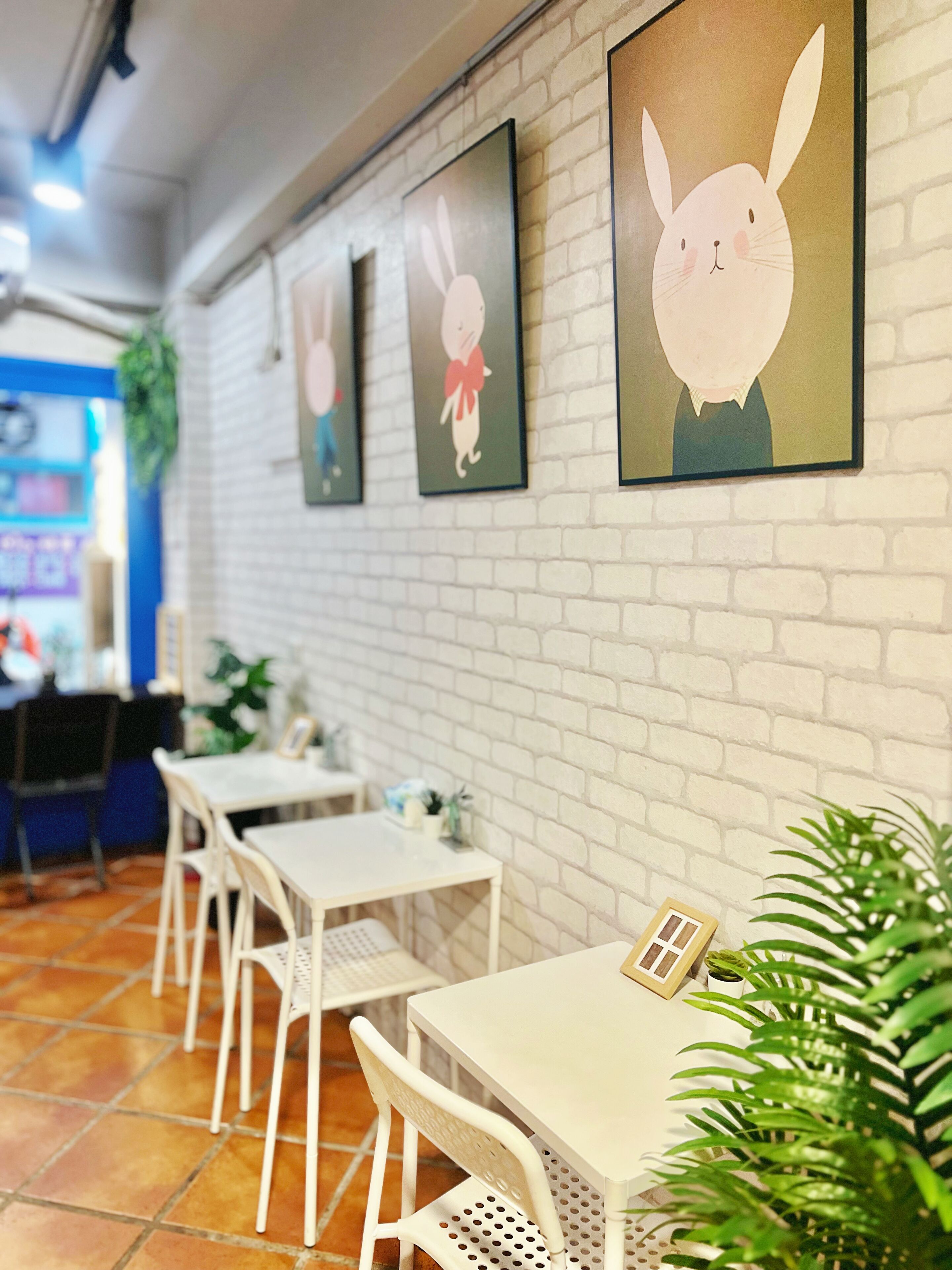 L’intérieur d’un restaurant présente des murs en briques blanches, des œuvres d’art encadrées, des tables et des chaises en métal blanc, ainsi que des plantes en pot.
