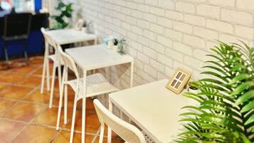 L’intérieur d’un restaurant présente des murs en briques blanches, des œuvres d’art encadrées, des tables et des chaises en métal blanc, ainsi que des plantes en pot.