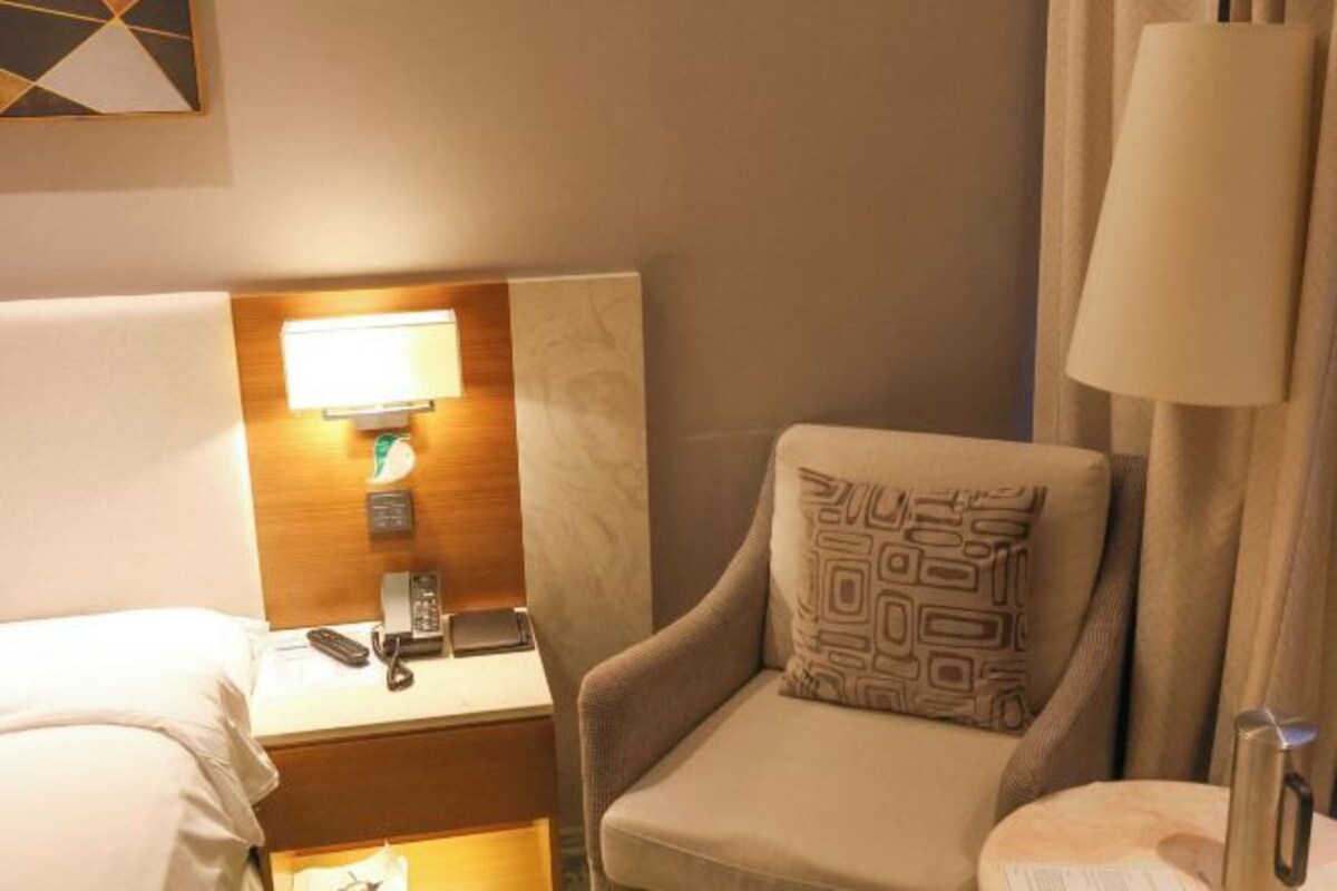 Une chambre d’hôtel comprenant un lit, une table de chevet avec une lampe, un téléphone, une chaise avec un coussin à motifs et un rideau.