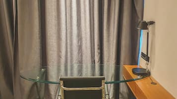 Una habitación con un escritorio de vidrio, una silla, una lámpara y una cortina.