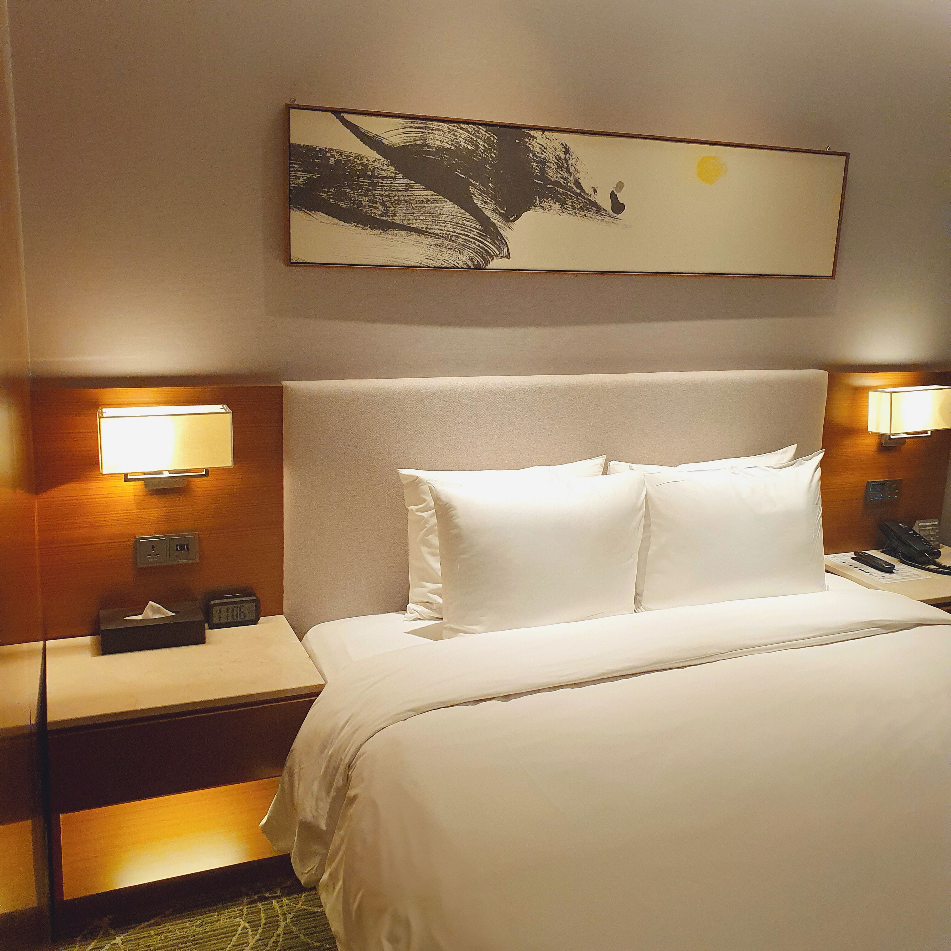 A hotel room with a large bed, two bedside lamps, a nightstand with a telephone, and a painting on the wall.
