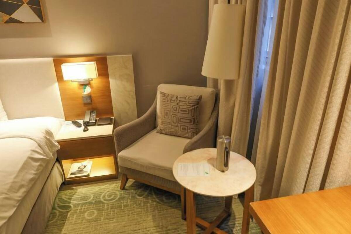 Une chambre d’hôtel avec un lit, une chaise dotée d’un coussin à motifs, une table de chevet avec une lampe, une petite table ronde avec une bouteille d’eau et un rideau.