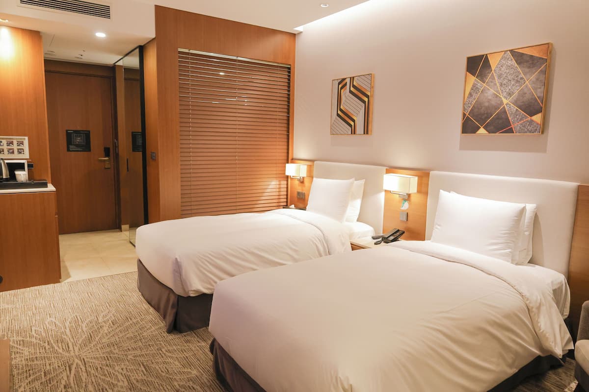 Une chambre d’hôtel avec deux lits, une porte en bois, une œuvre d’art accrochée au mur et un bureau avec une lampe.