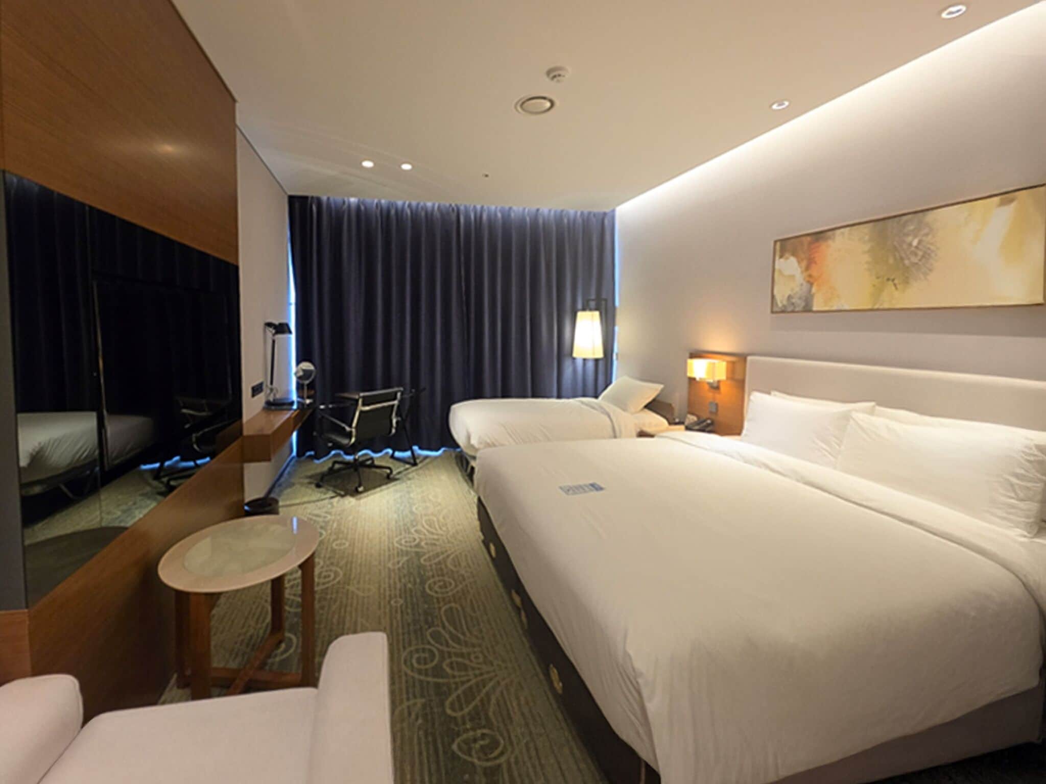 Habitación de hotel con dos camas, un escritorio, una silla, una mesita, una lámpara y un cuadro en la pared.